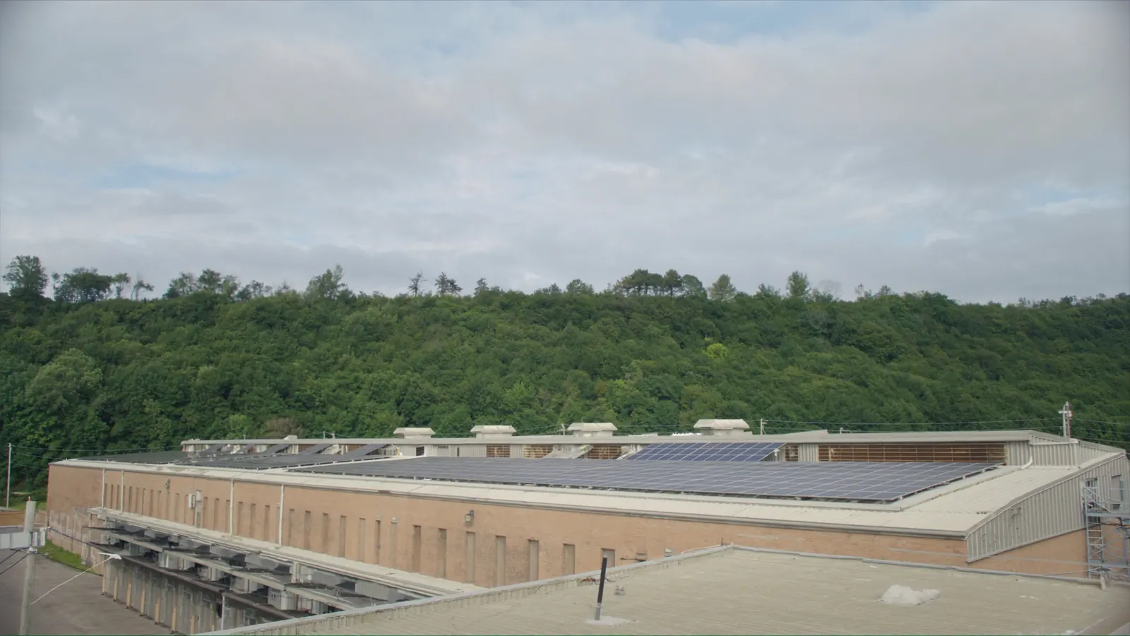 A brick light-industrial building with solar panels on its roof and the wooded escarpment behind it