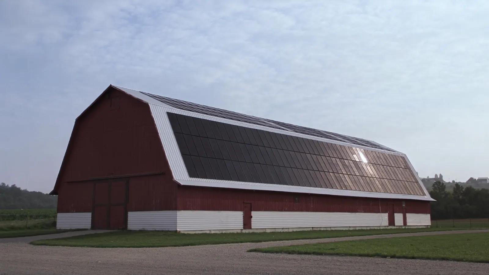 A long red dairy barn whose whole south roof is covered in black solar panels, farmland in front